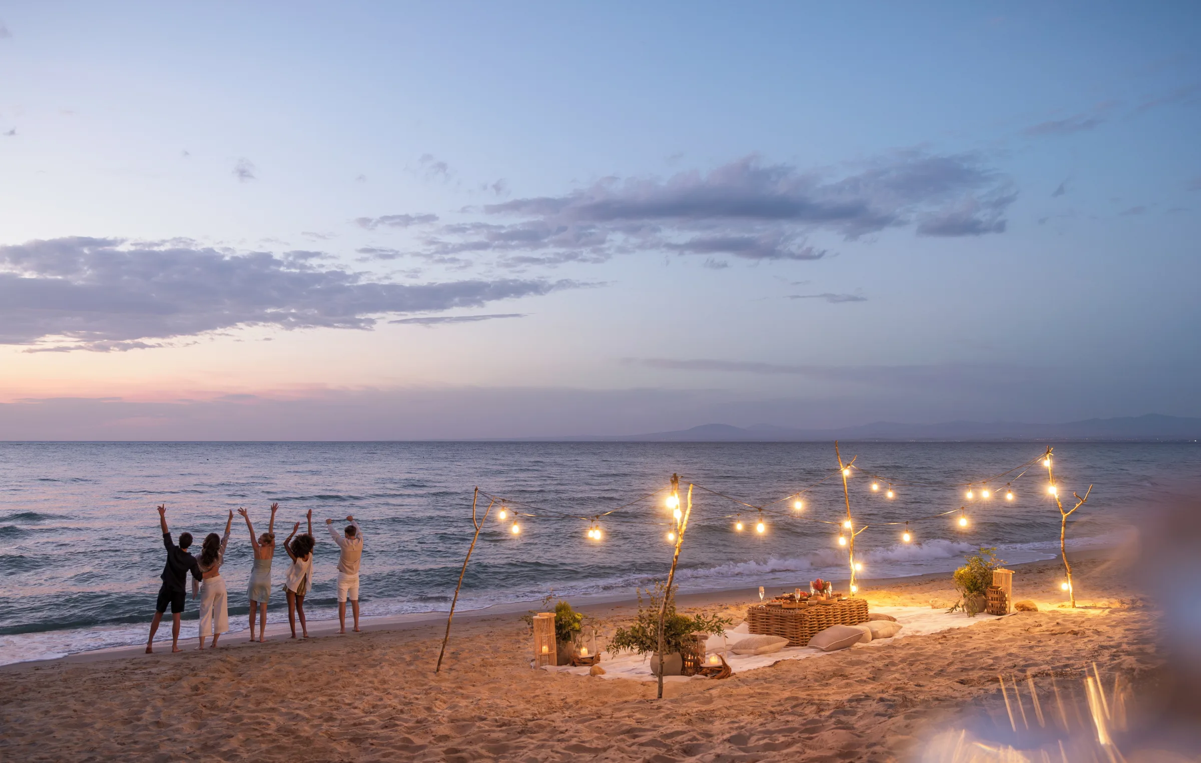 Friends gathering on a beach beside a candlelit table at dusk