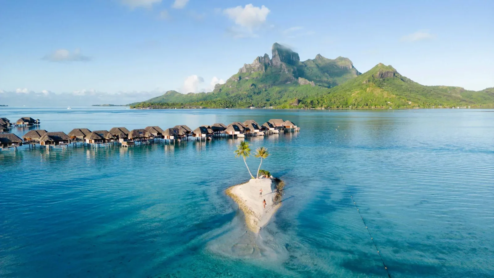 Overwater bungalows and a palm-fringed islet in a turquoise Polynesian lagoon beneath green mountains